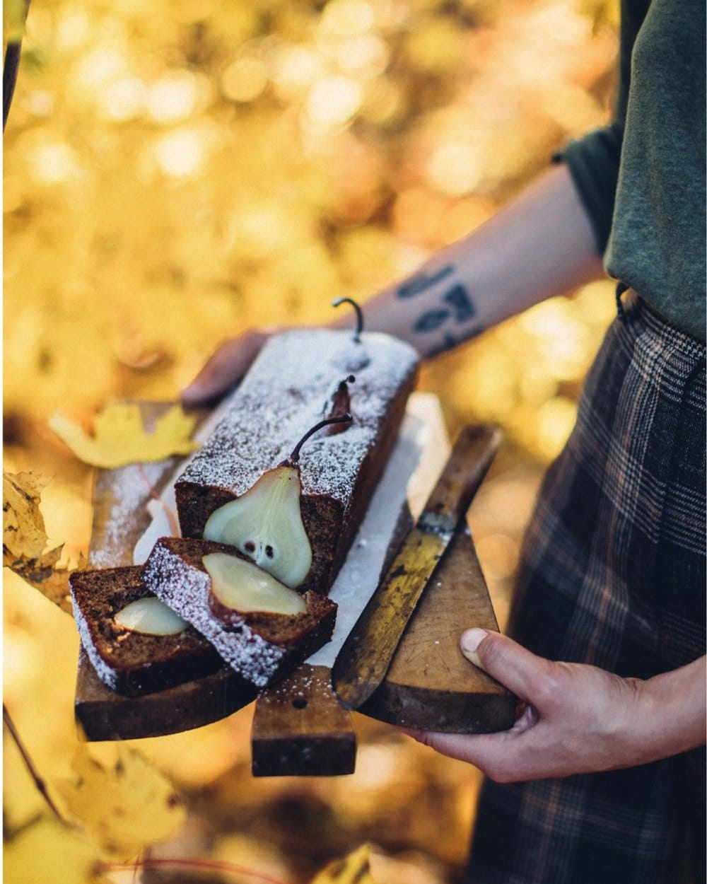 Slices of pear and walnut loaf cake dusted with icing sugar, with whole baked pears inside, held on a wooden board against golden autumn leaves