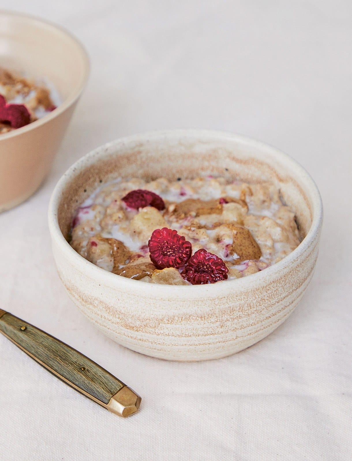 A ceramic bowl of creamy porridge with diced pear, almond butter and raspberries, beside a wooden-handled knife