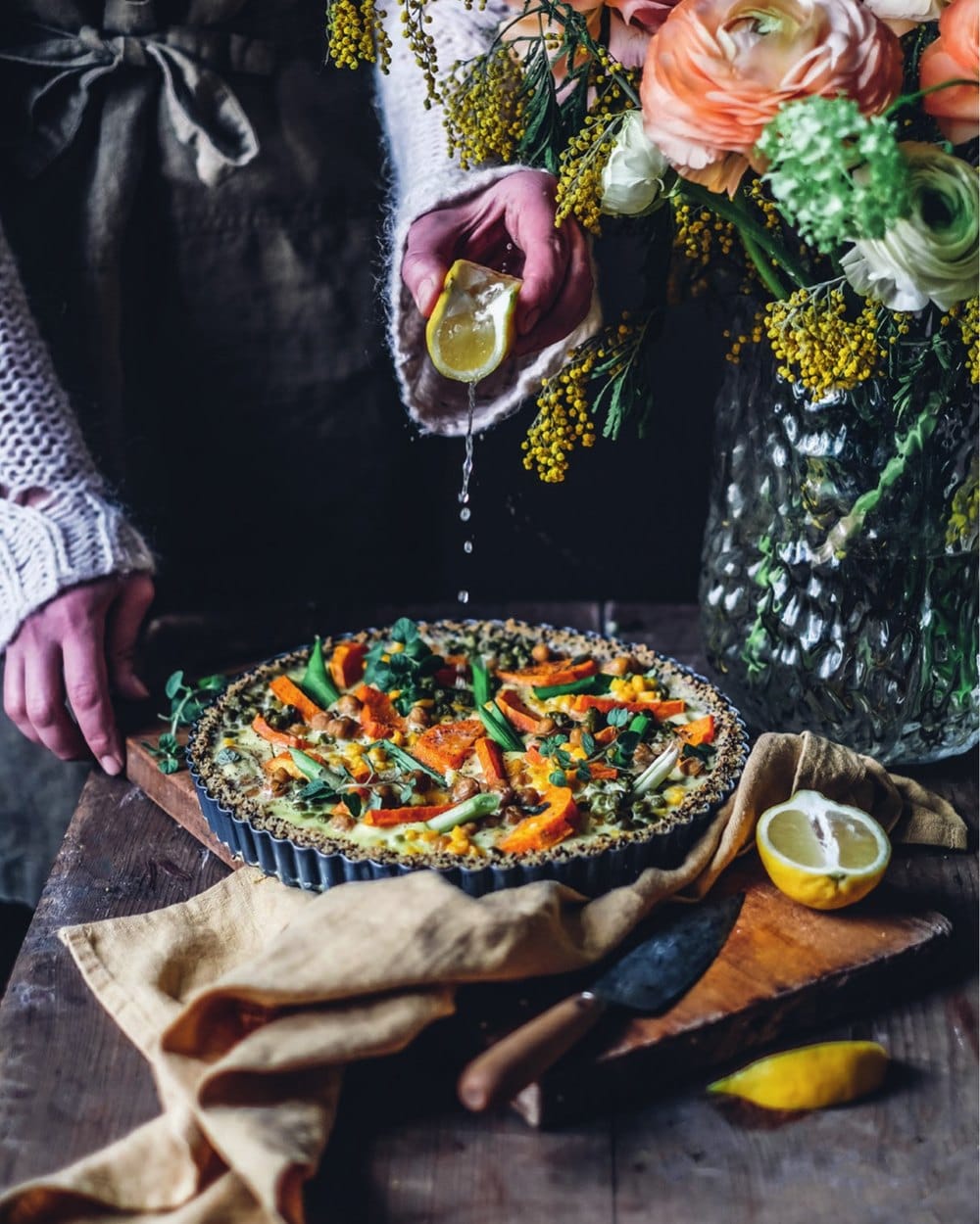 A sweet potato, chickpea and pea quiche in a fluted tart tin on a wooden board, with a hand squeezing lemon over it