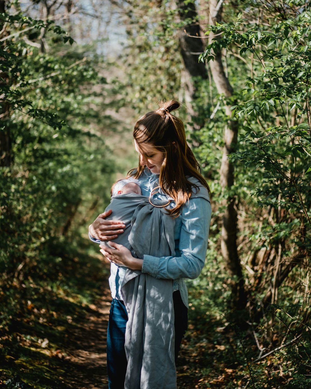Photograph of a mother standing in sun dappled woods with her baby worn against her chest in a ring sling