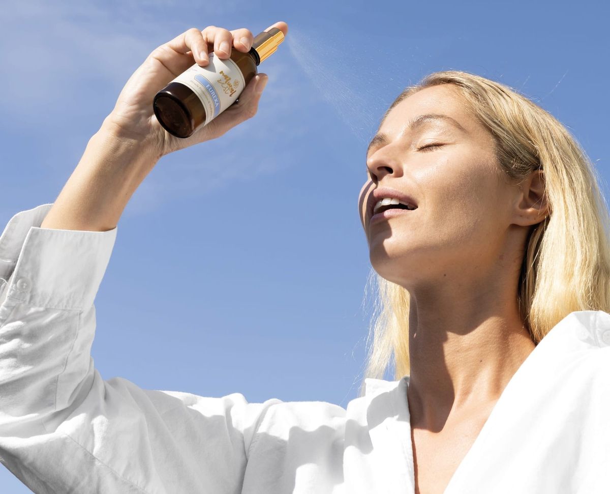Woman with closed eyes holding up a dropper bottle of skincare oil against a blue sky