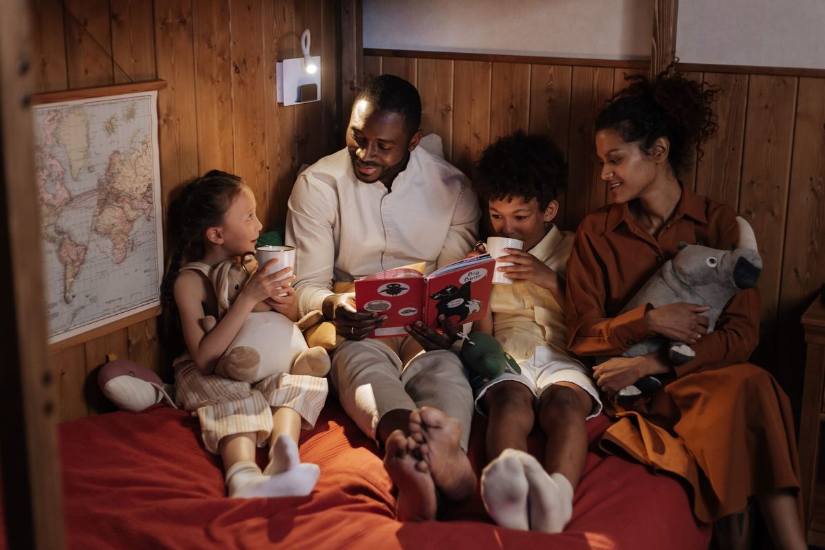 A family curled up together on a bed in a cosy wooden room, sharing a picture book with mugs and cuddly toys
