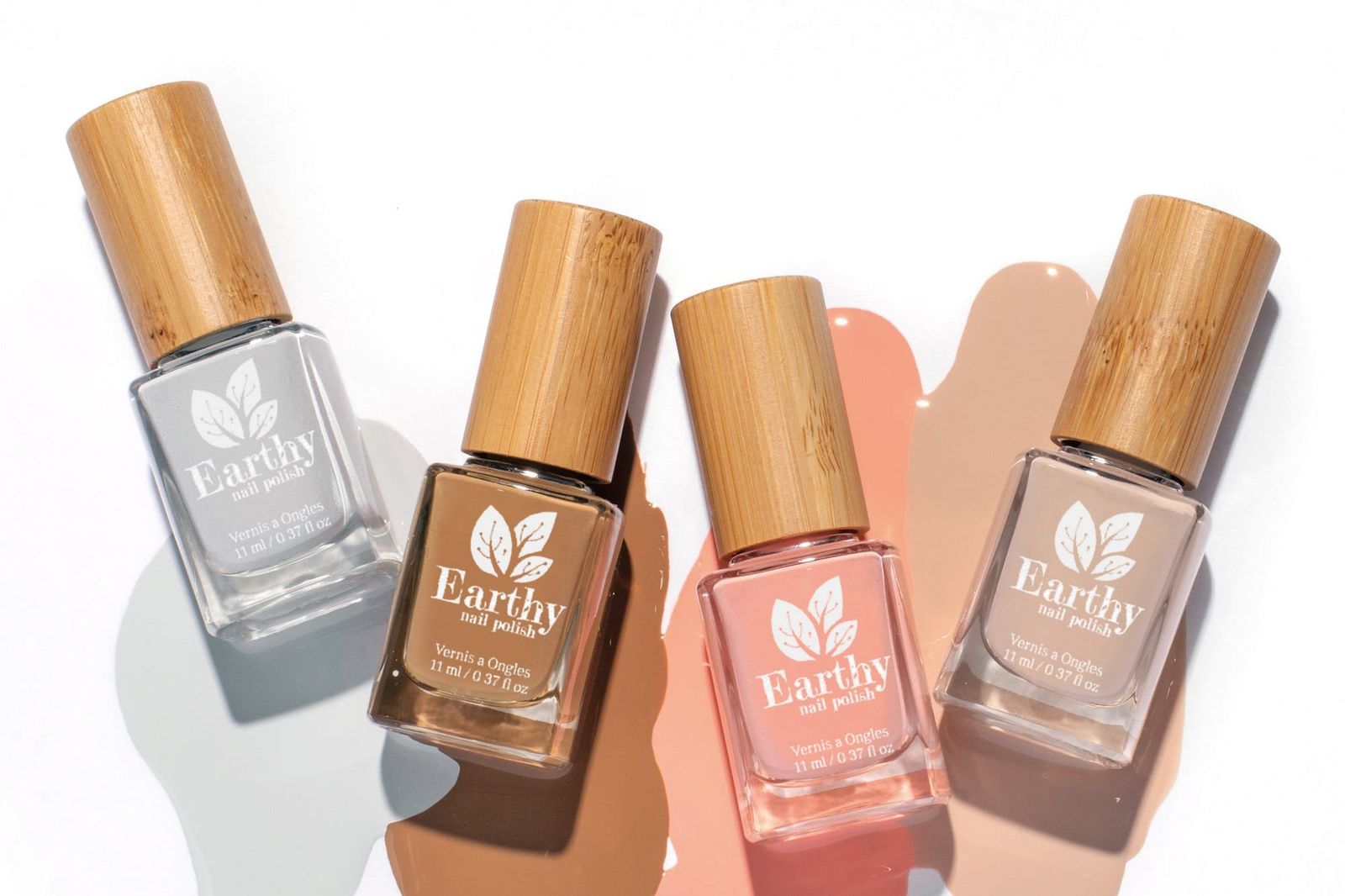 Four Earthy nail polishes in grey, chocolate brown, coral pink and nude, lying across matching spills of polish on a white background