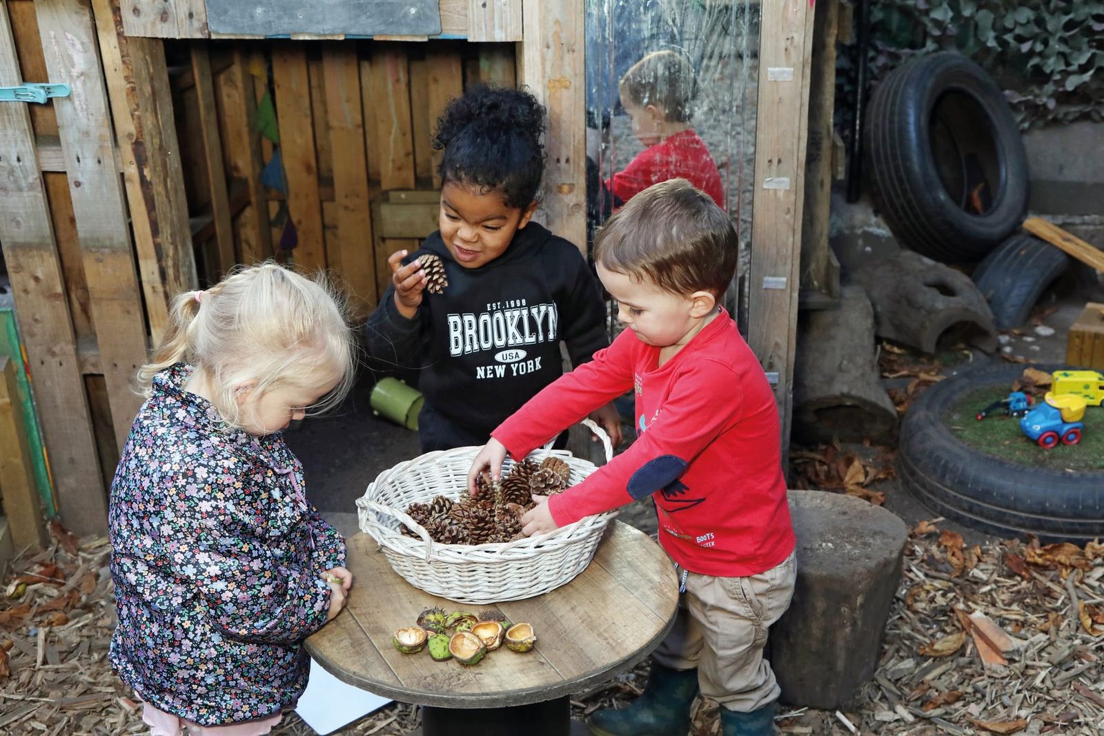 Three young children gathered around a small log table with a basket of pine cones and chestnut cases in a wooden outdoor play area