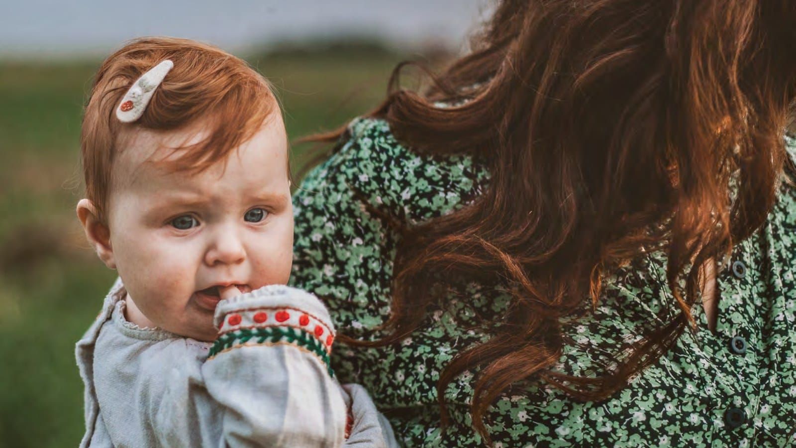 A baby with red hair held on her mother’s shoulder in a green field