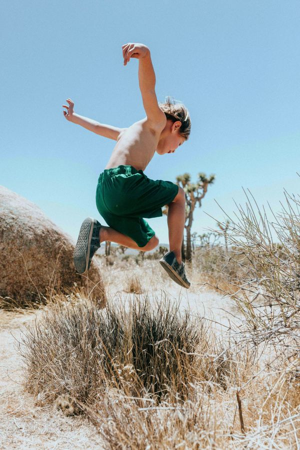 Child in shorts leaps over a patch of grass in a rocky, sandy landscape