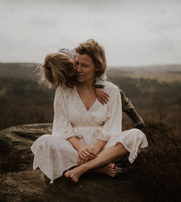 Photograph of a barefoot mother sitting on a rock in a white dress, with her son draped around her shoulders