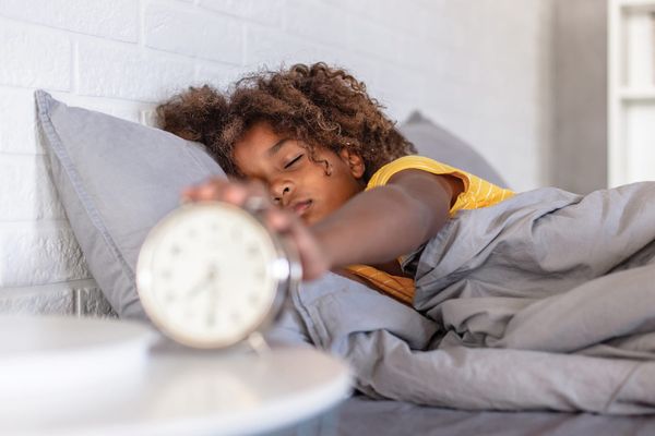 A sleepy girl with curly hair reaching out from under a grey duvet to switch off an alarm clock on her bedside table