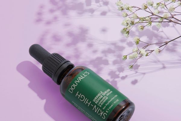 A bottle of Douvall's Skin High wrinkle rehab serum lying on a lilac background with the shadow of gypsophila flowers
