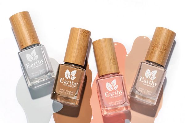 Four Earthy nail polishes in grey, chocolate brown, coral pink and nude, lying across matching spills of polish on a white background