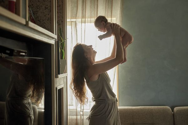 A mother lifting her baby up high in a sunlit living room, light streaming through the curtains