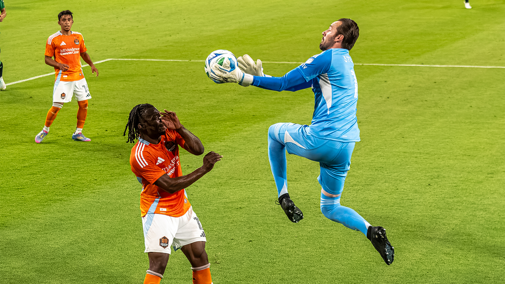 Brad Stuver leaping and catching ball in front of Houston Dynamo player