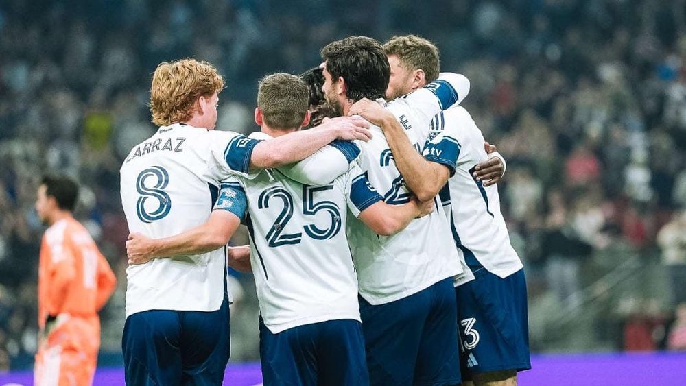 A group of Vancouver Whitecaps players celebrating on field with an upset goalkeeper in the background