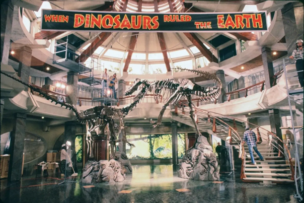 A production still from Jurassic Park (1993) showing the set for the interior of the visitors’ center. The interior is a rotunda featuring green marble floors; a curved, concrete staircase with red steel balusters; a mezzanine overlooking the first level; and concentric coffered windows decreasing in size as they approach the oculus.