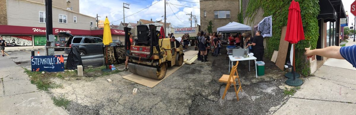 A steamroller is set to drive across an extra large relief carving to print image on fabric. People are engaged in activities around the lot.