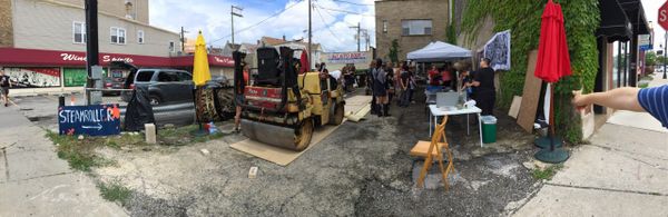A steamroller is set to drive across an extra large relief carving to print image on fabric. People are engaged in activities around the lot.