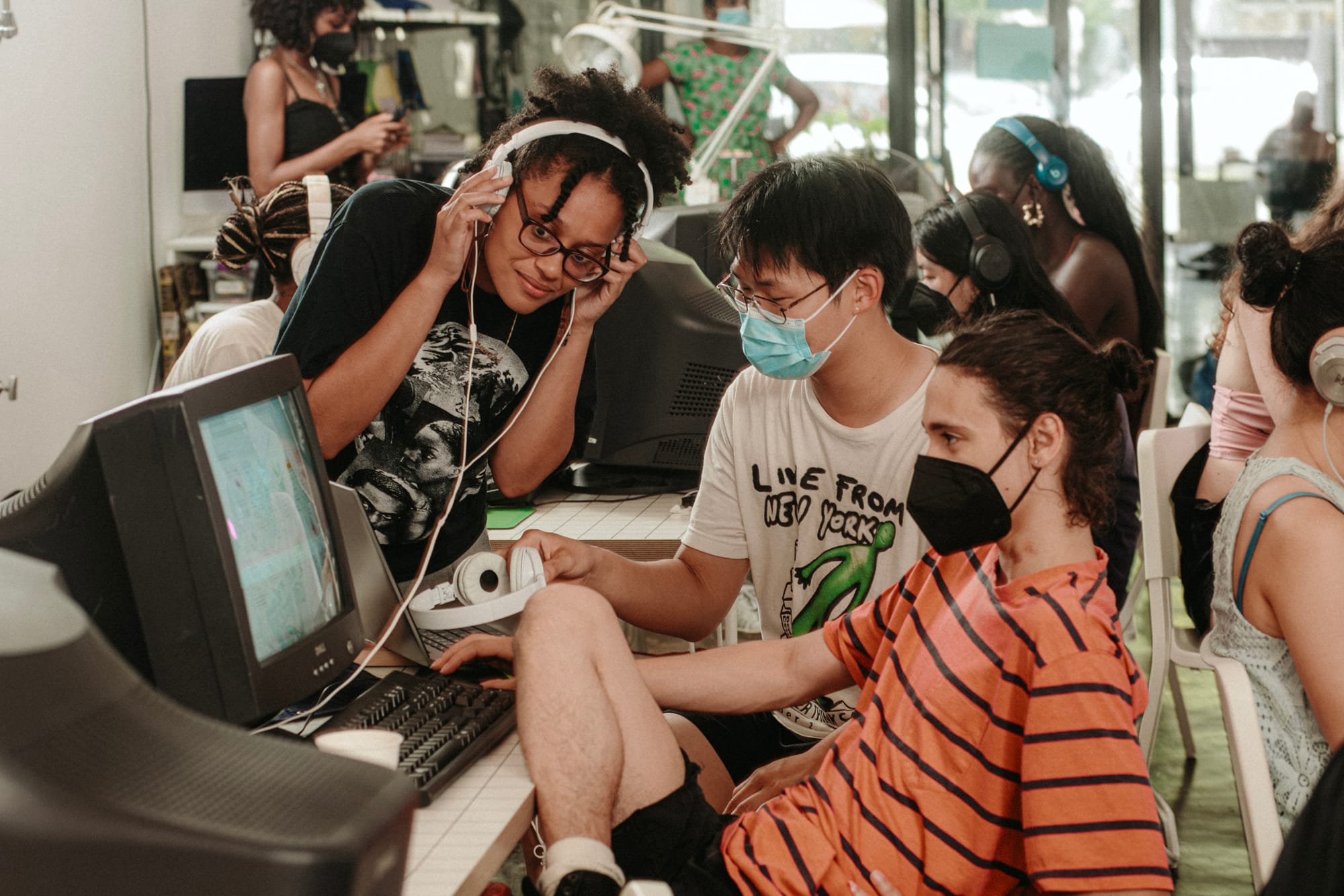 Readymag blog: A group of young people in a retro computing space; three individuals wearing masks and headphones look together at a CRT monitor while handling controls and listening to audio, surrounded by more participants working on old computers.