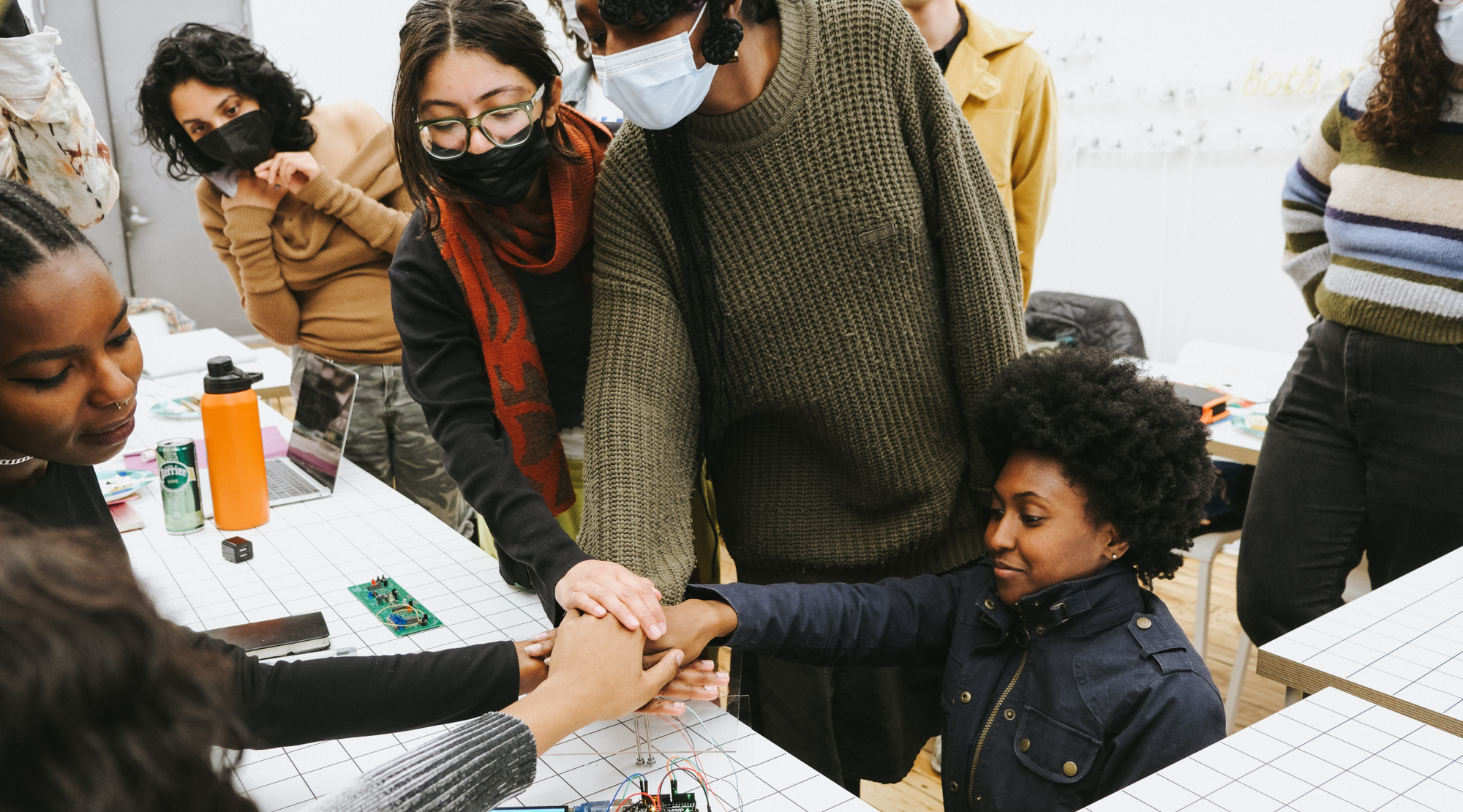Readymag blog: A group of people gather around a table covered with a white grid-patterned surface, extending their hands into a communal stack over a small electronics setup with wires and a microcontroller. 