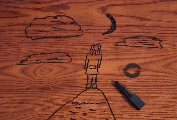A drawing on wood of a person standing on a mountaintop, looking at a crescent moon and clouds. There is a black kissprint on the right, with an open tube of black lipstick sitting underneath