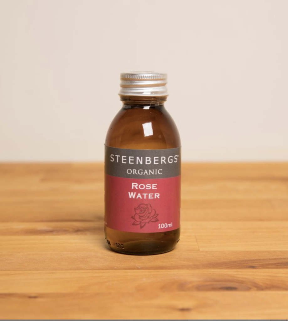 A 100ml brown bottle with a red, grey, and white label reading 'Steenbergs' Organic Rose Water' is contained by a silver twist cap. The bottle sits on a light-coloured wooden table with a grey wall behind it.
