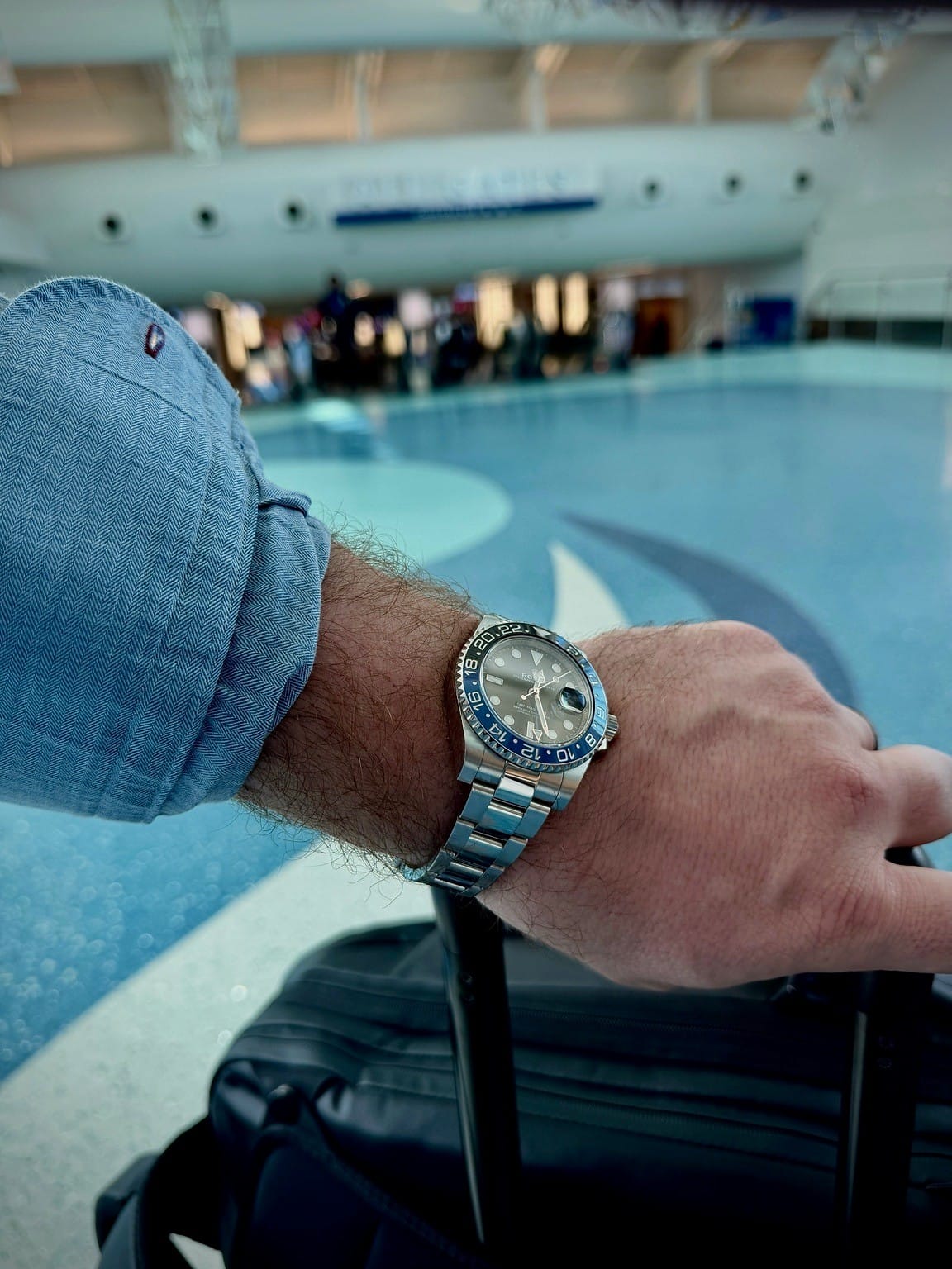 Rolex GMT-Master II on a wrist beside a blue shirt cuff and suitcase handle in an airport terminal.