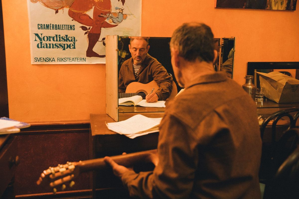 Jonathan Richman is sitting a a desk with his guitar, his back to the camera, writing in a notebook. 