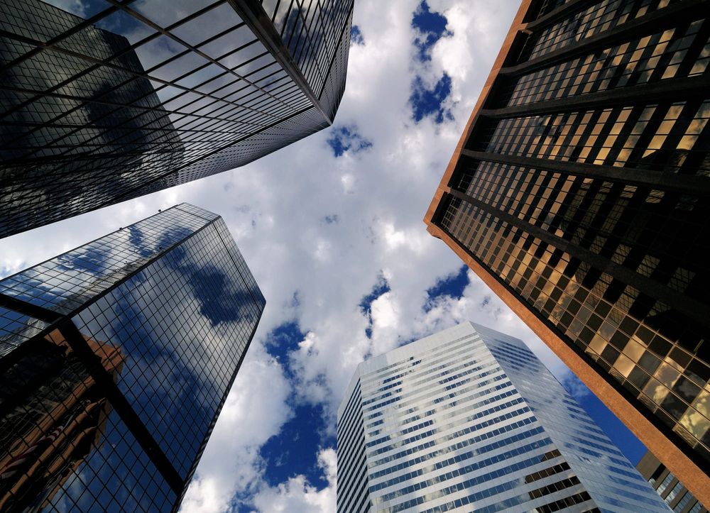Looking up at a cluster of downtown office towers with glass facades against a blue, cloud-filled sky