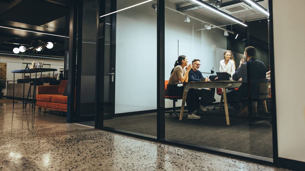 Four coworkers meeting in a glass-walled conference room inside a small modern office suite