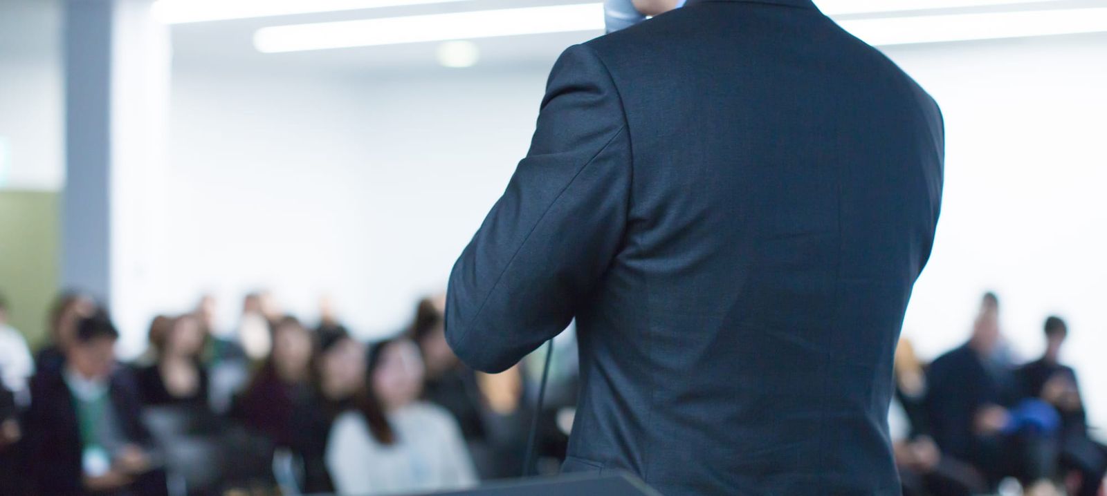 Speaker in a dark suit addressing a seated audience at a business conference