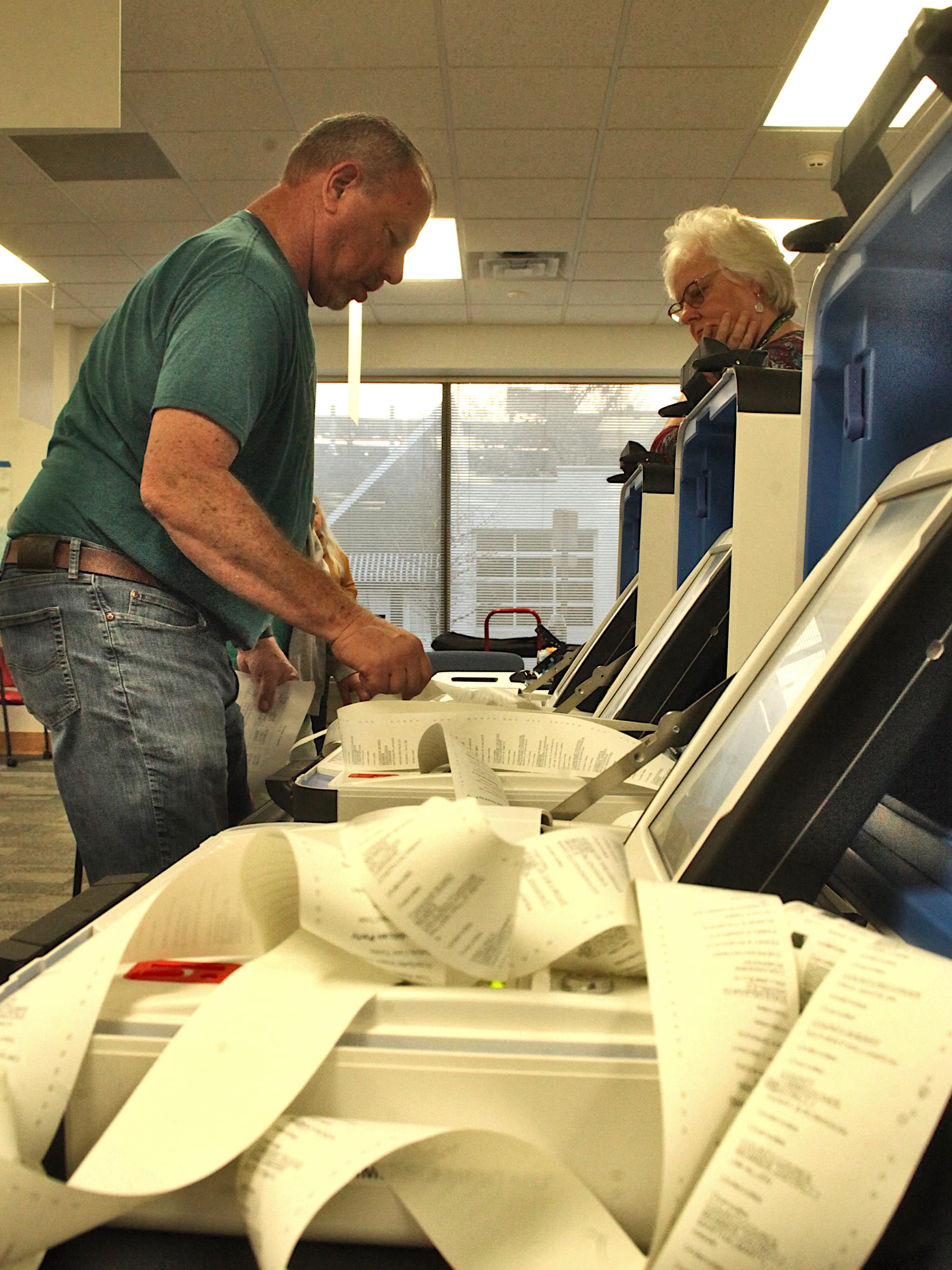 man standing to left of voting machines with scrolls of paper