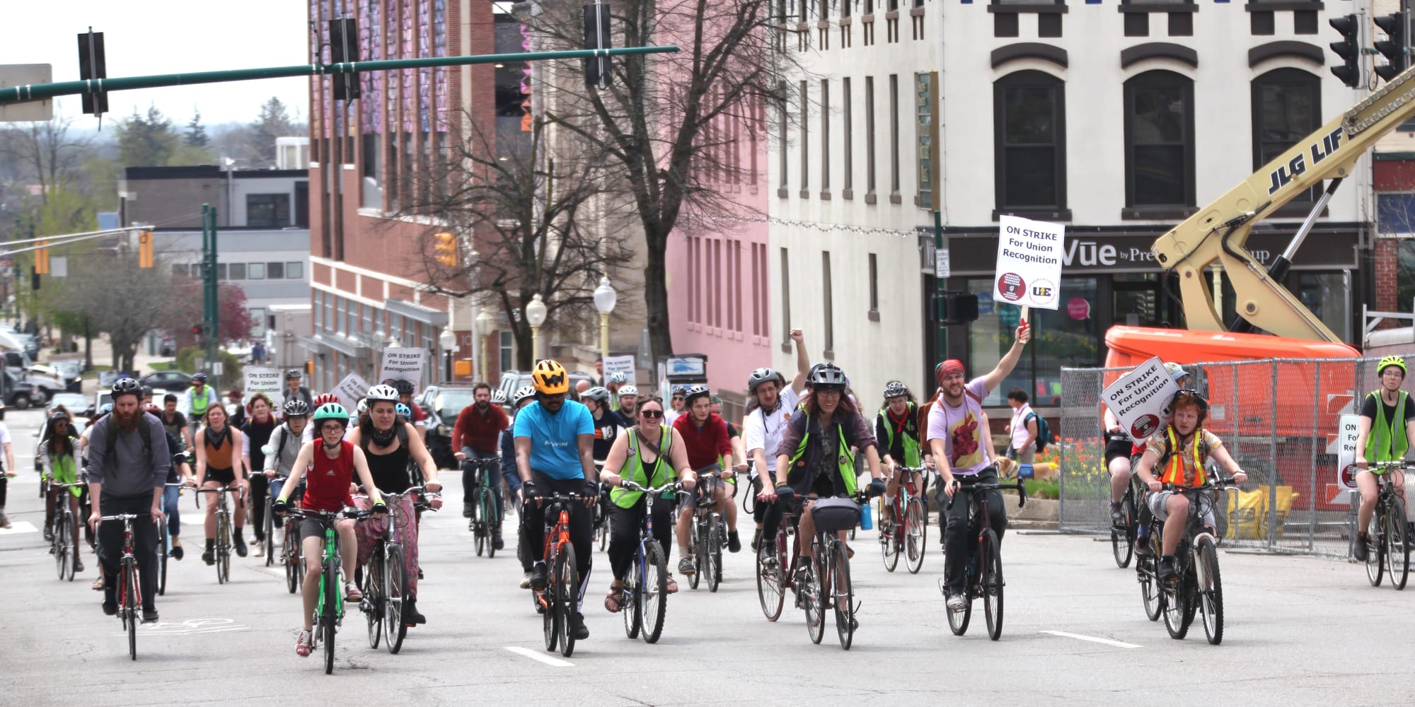 group of bicyclists riding up the street taking up the full width of the street holding signs that say, "ON STRIKE for union recognition"