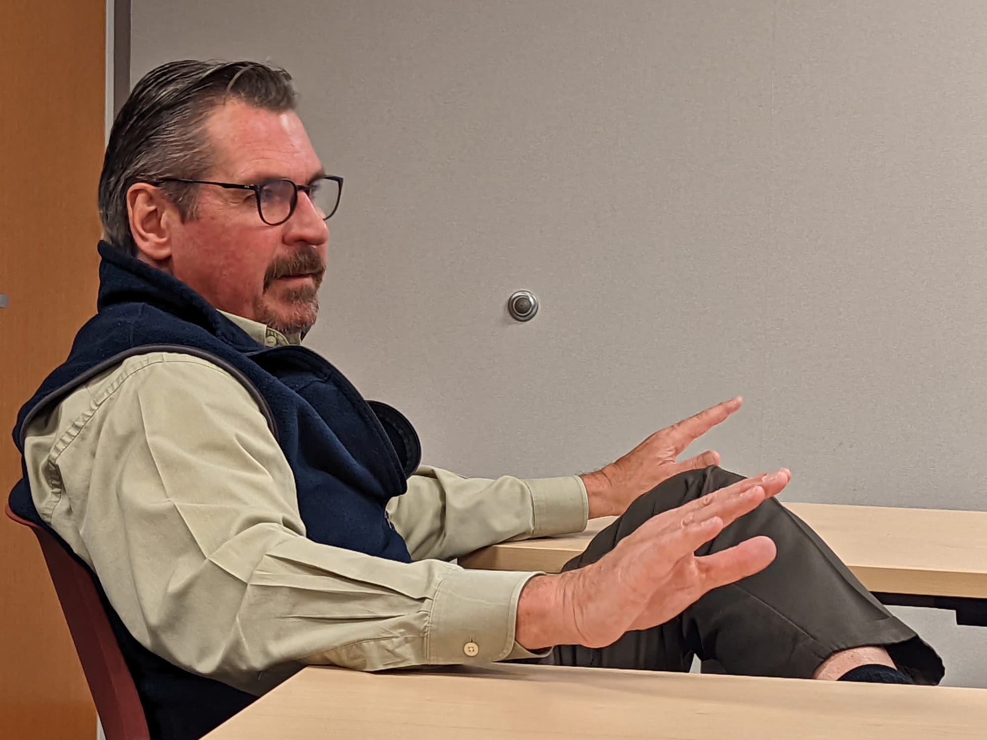 man seated in chair oriented to the right speaking and gesturing