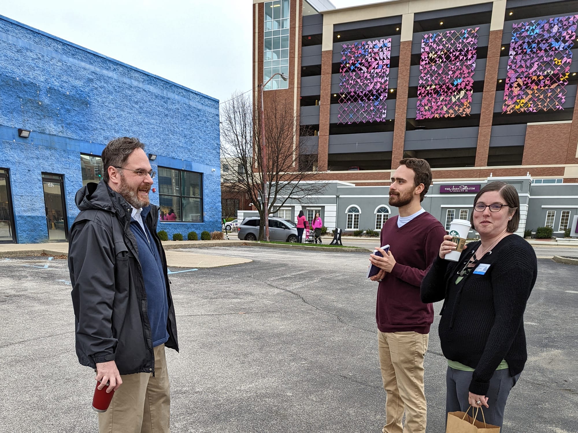 view of blue building on the left parking lot in foreground, with three people standing talking together,and parking garage with colorful tiles in the background to the right