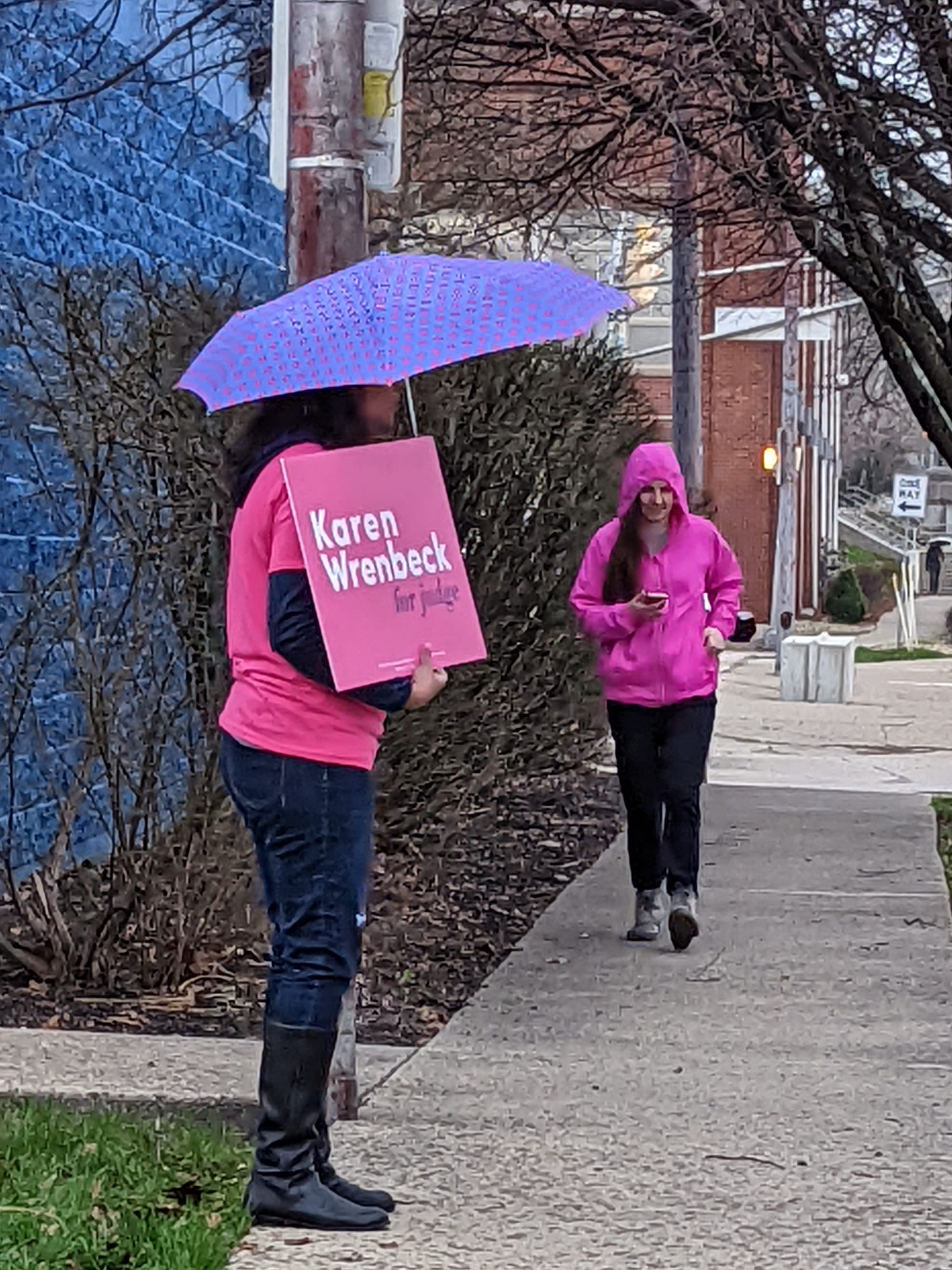 campaign volunteer holding a sign that says "Karen Wrenbeck" wearing a pink shirt that matches the sign and a woman father down the sidewalk wearing a pink sweatshirt