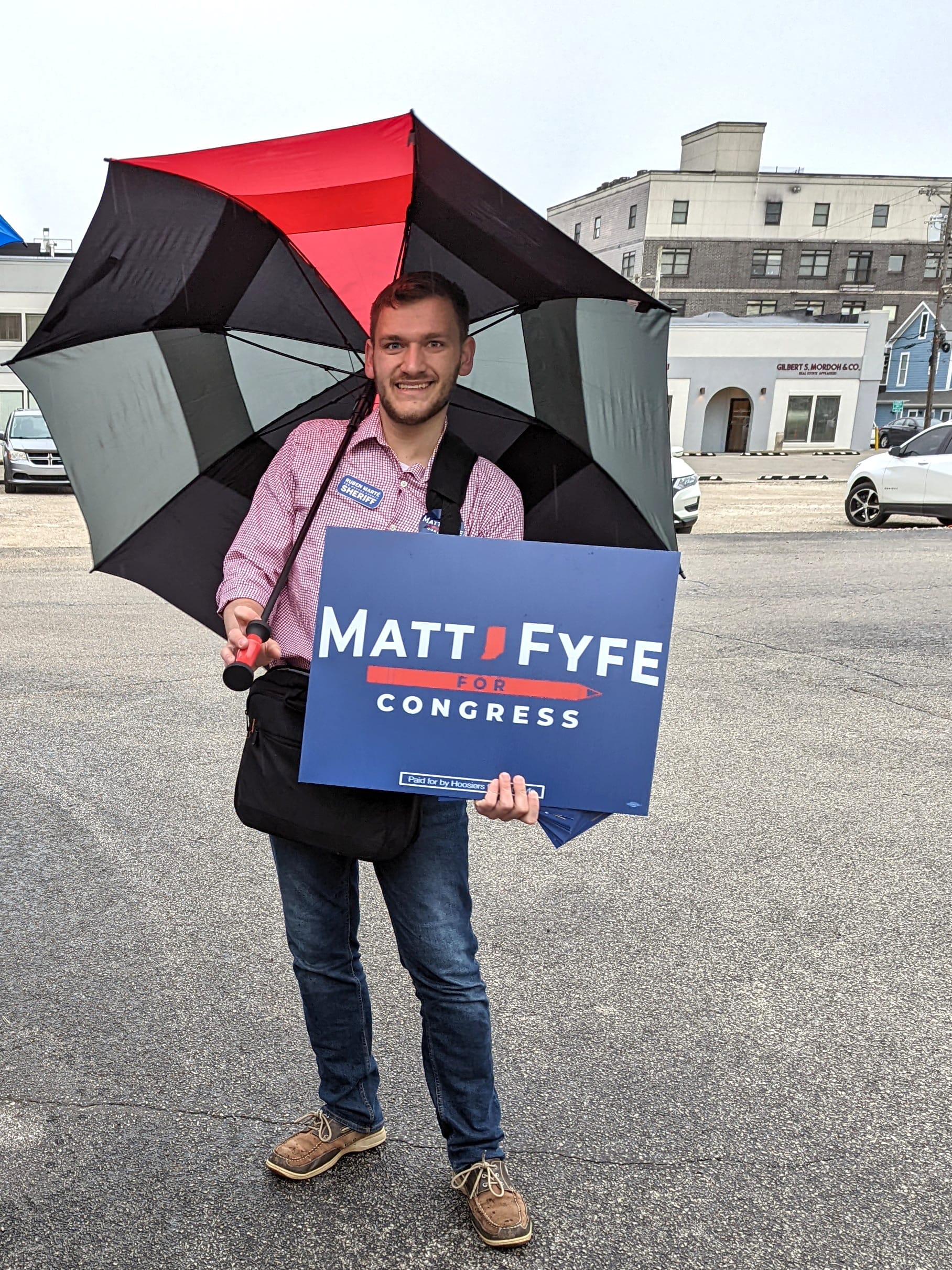 man holding an umbrella in his right hand and a blue campaign sign in his left hand posing for the camera
