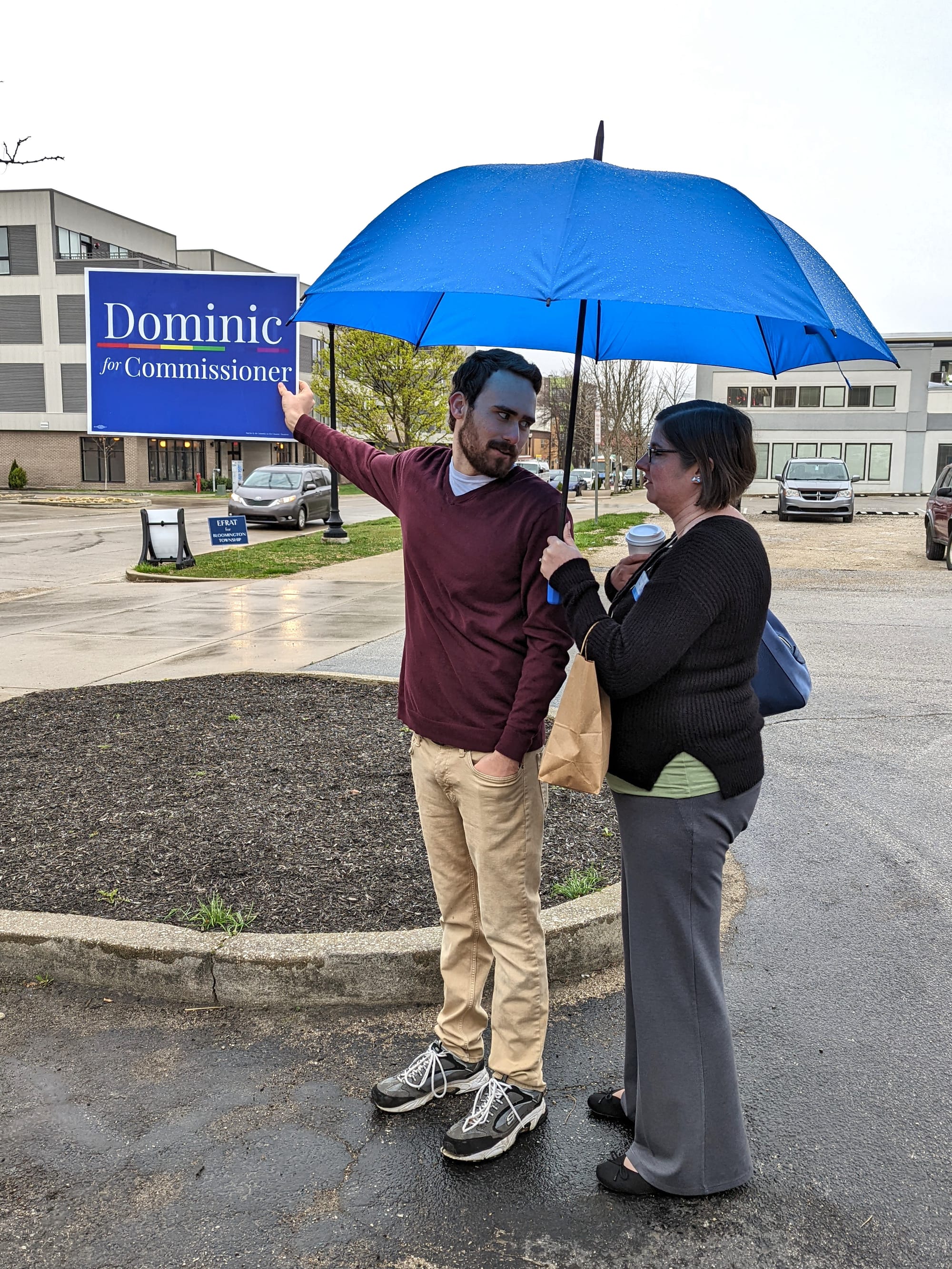 Man standing under and umbrella held by a woman. The man is holding a campaign sign with an extended arm, looking back over his shoulder at the woman
