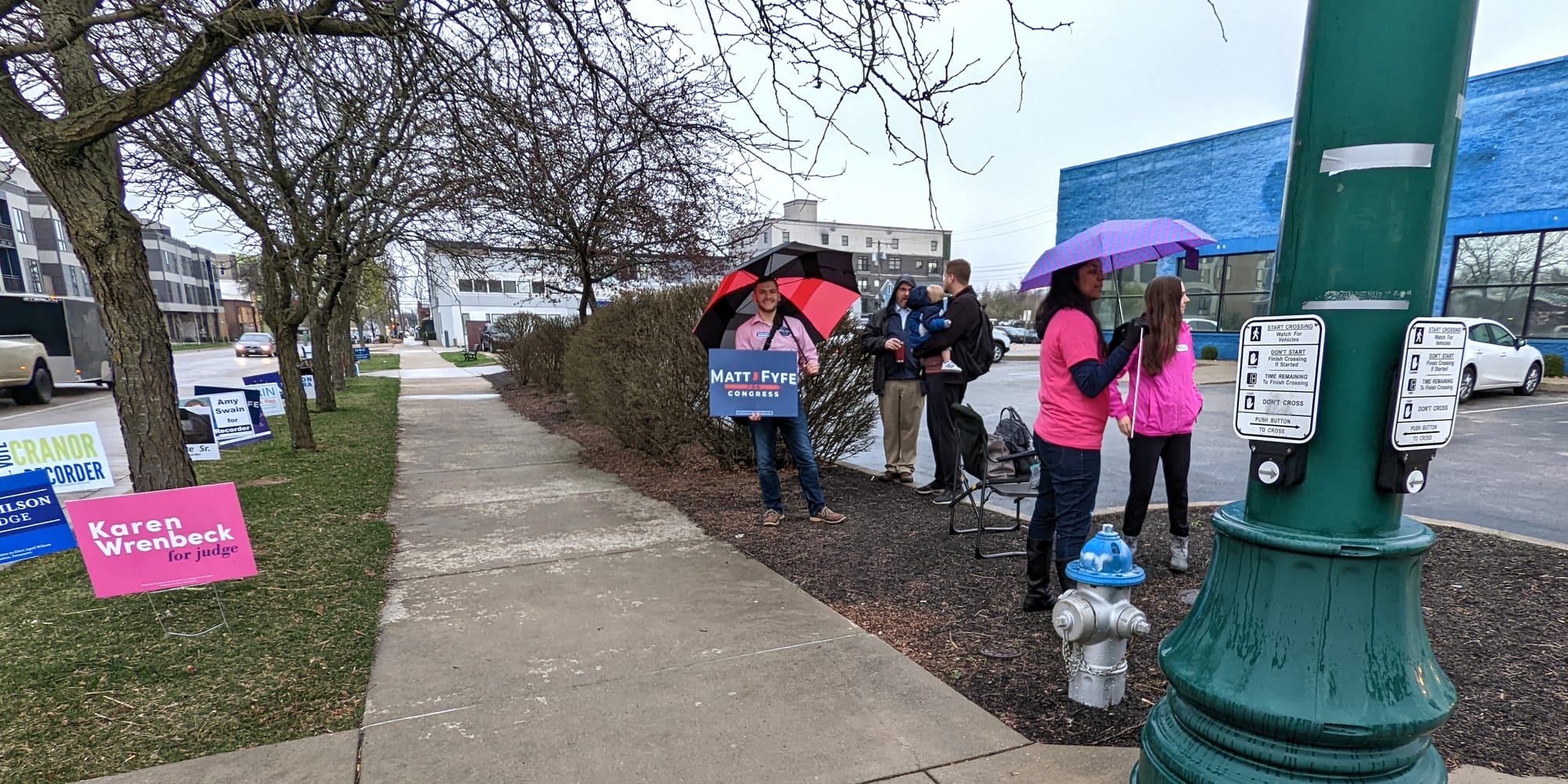 view down sidewalk with campaign signs on one side and candidates holding signs on the other