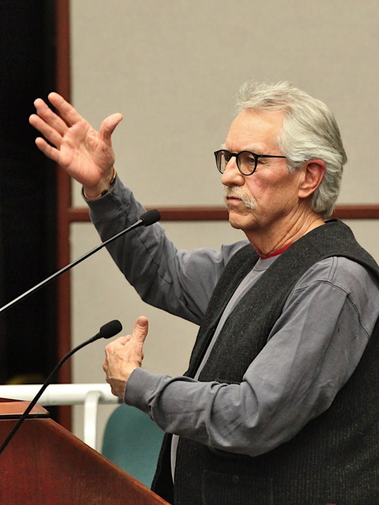 man with glasses speaking at podium gesturing with his right hand