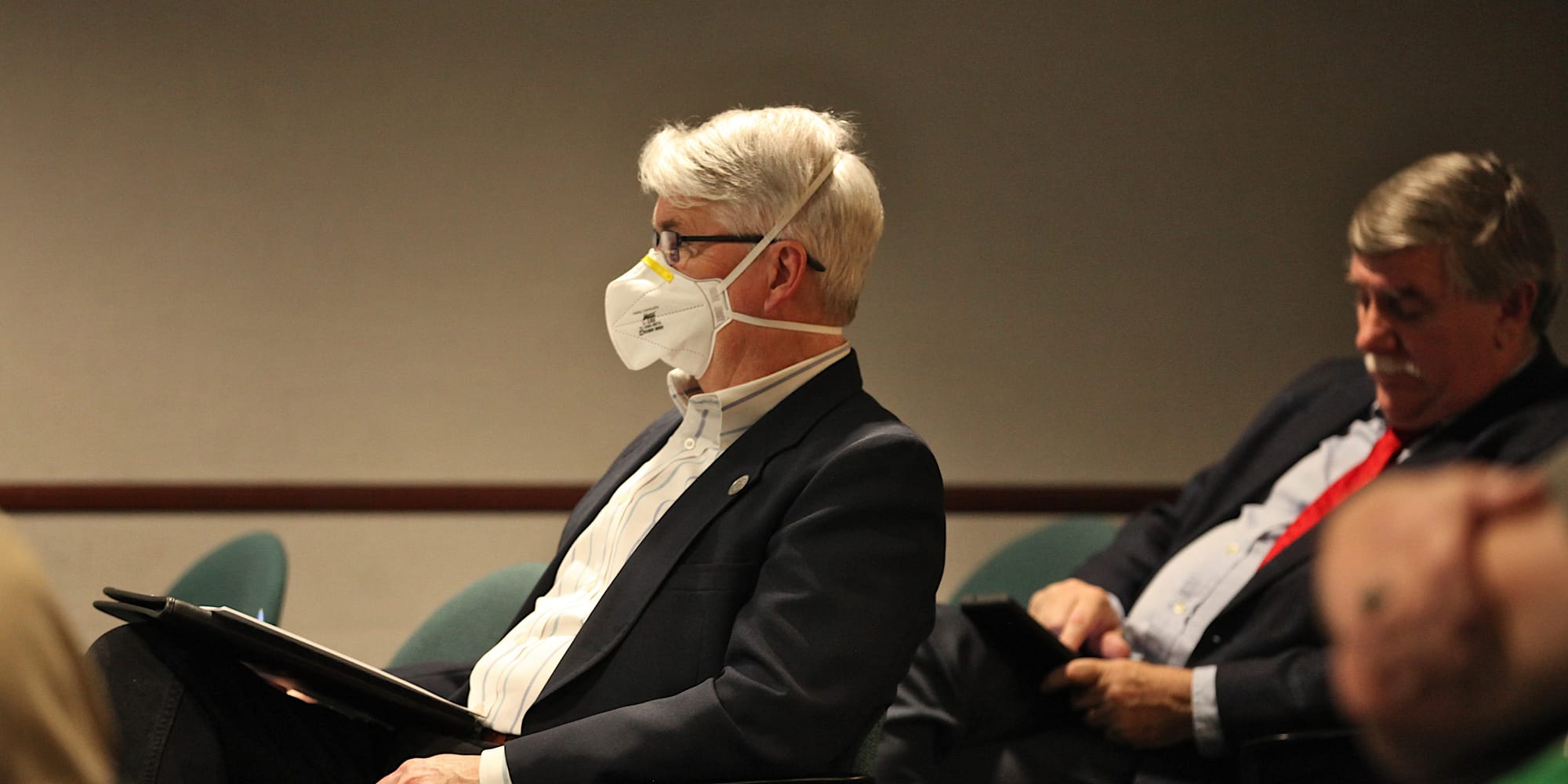 man wearing mask seated in city council chambers looking to the left