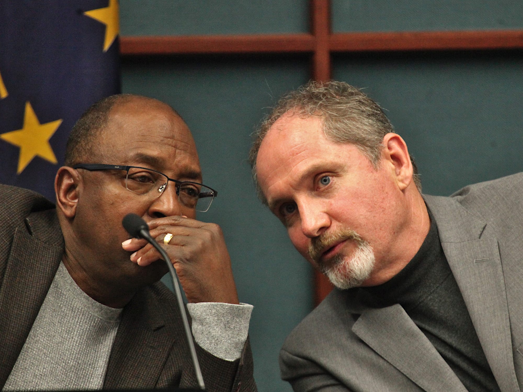 two men seated at council dais leaning towards each other talking