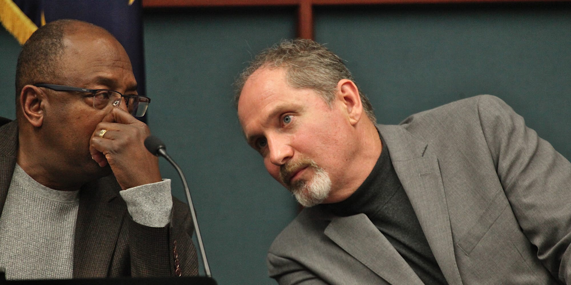two men seated at council dais leaning towards each other talking