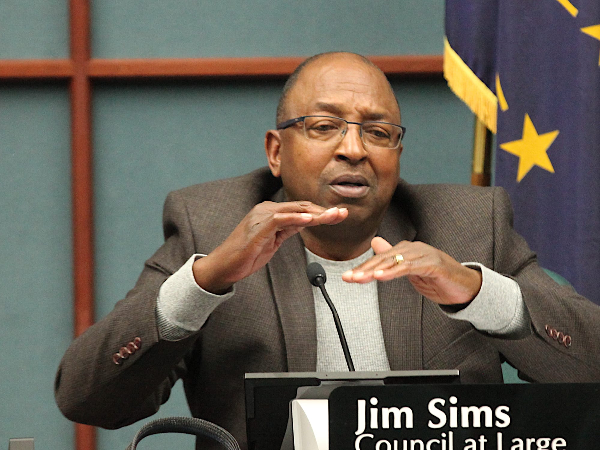 man sitting at city council table with name plate mic holding his hands flat a certain distance apart vertically