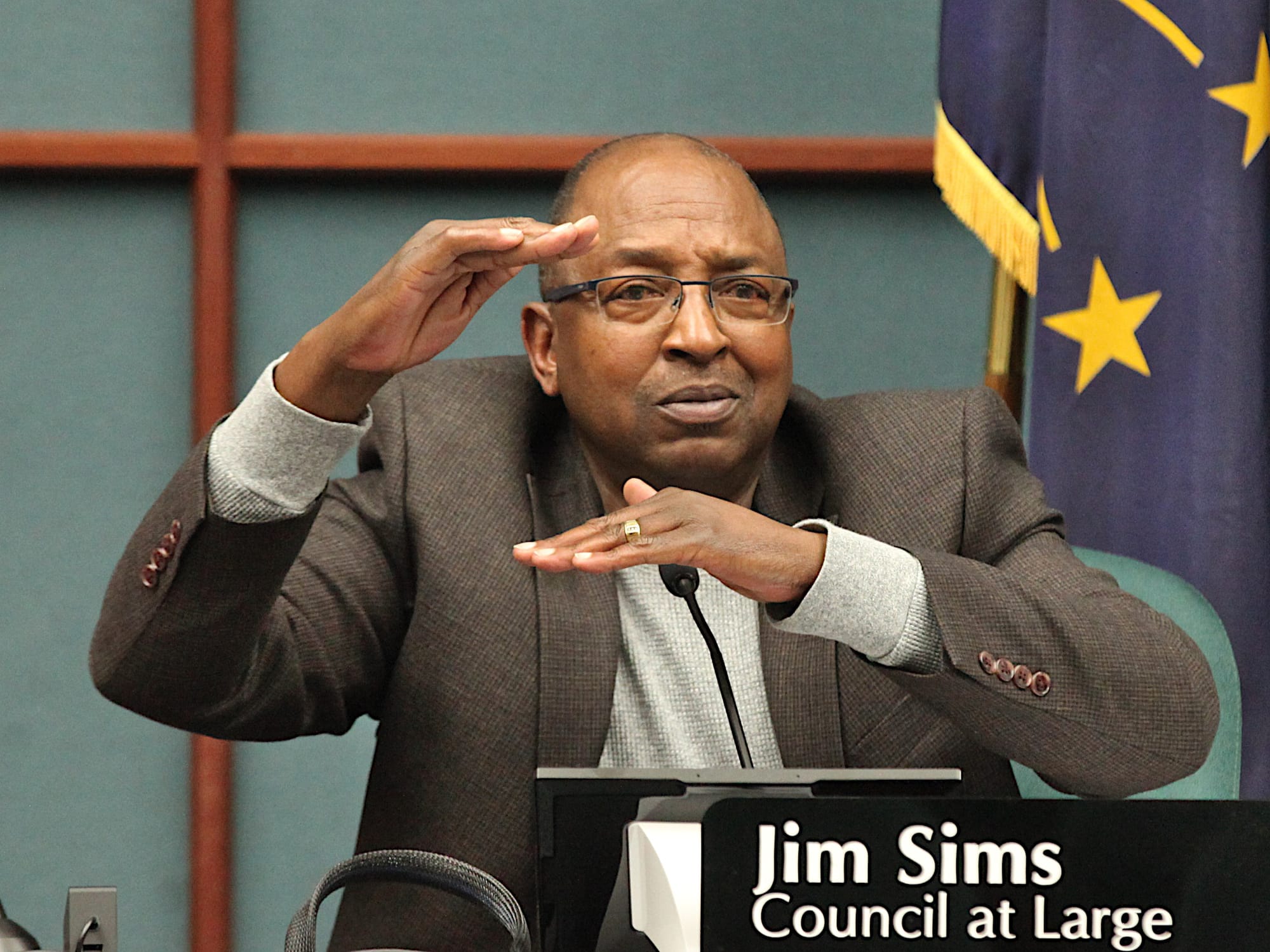 man sitting at city council table with name plate mic holding his hands flat a certain distance apart vertically