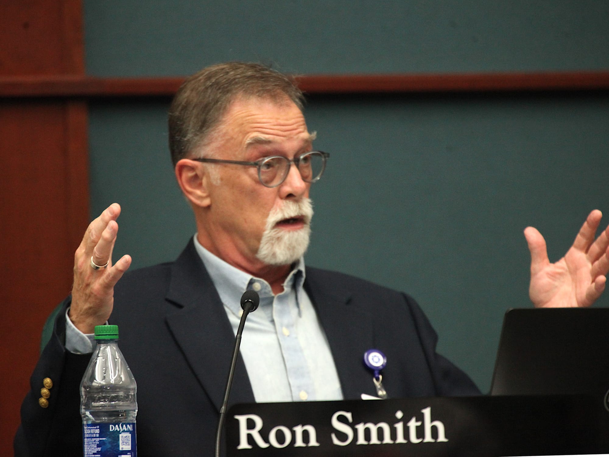 man sitting at city council table with name plate mic holding his hands flat a certain distance apart side to side