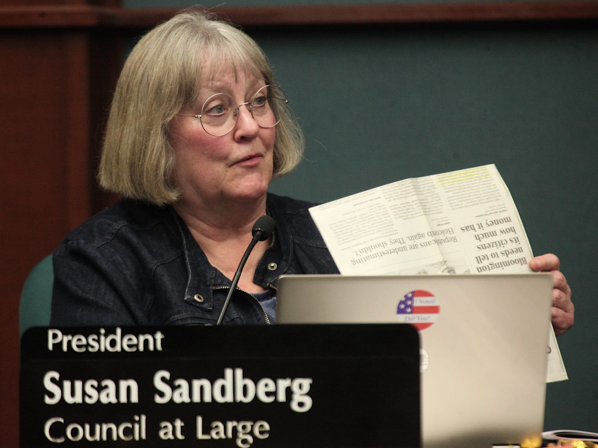 woman seated at council dais holding up a sheet of paper with a newspaper article printed on it