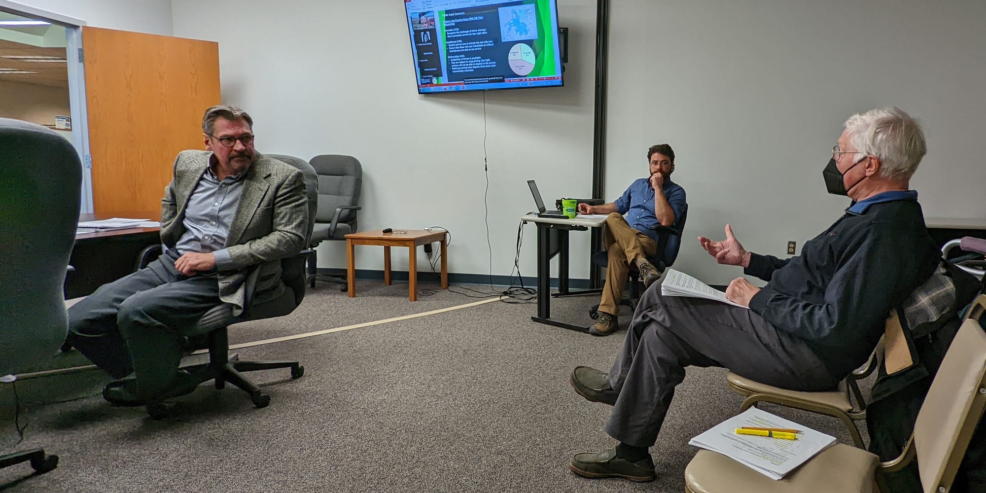 view of a board room with a man turning around to look at someone seated in the audience. Seated in the middle of the frame, but in the background is another man. A screen hangs on the wall.