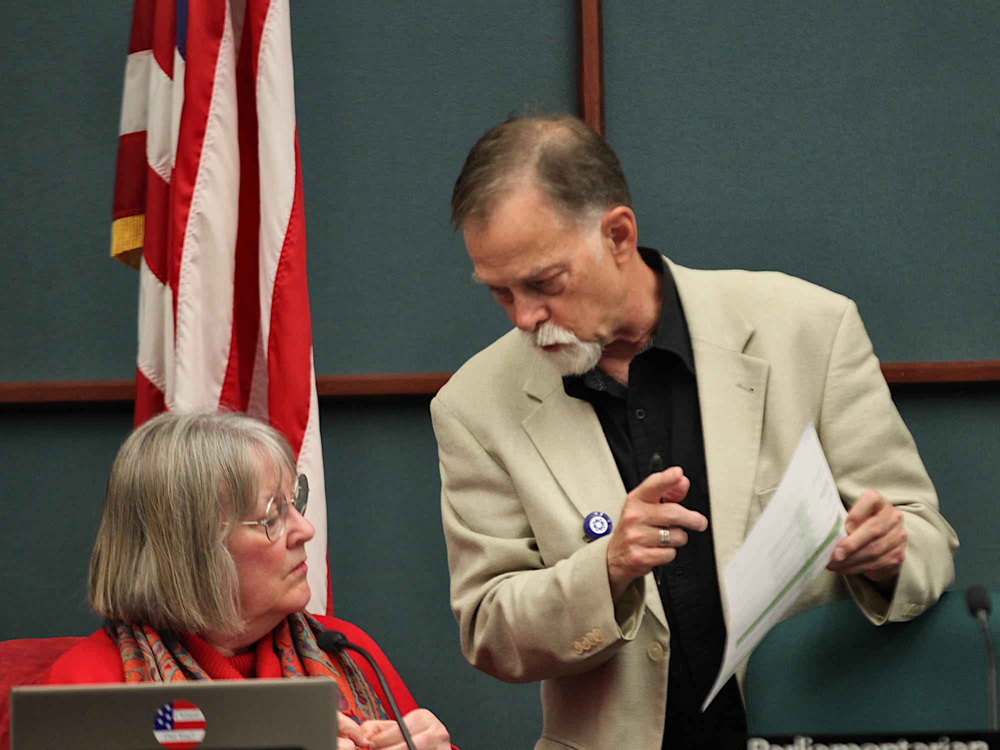 man standing showing sheaf of papers to a woman, who is seated