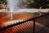 View of a city park and spray fountain as a boy runs along its edge. A low, chain link fence appears in the foreground with tables and benches in the background near a jungle gym.