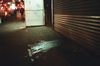 Nighttime shot of a city sidewalk with street lights, traffic, and people walking in the distance. On the right is a closed pull-down, corrugated storefront door next to a dirty sidewalk.
