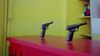 Closeup of a carnival table with fixed toy pistols aimed toward a wall. Colorful prizes appear on a shelf in the upper left.